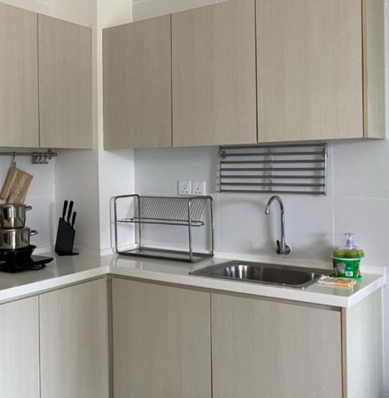 A modern kitchen with light-colored cabinets, a stainless steel sink, a kettle, and a green soap dispenser.