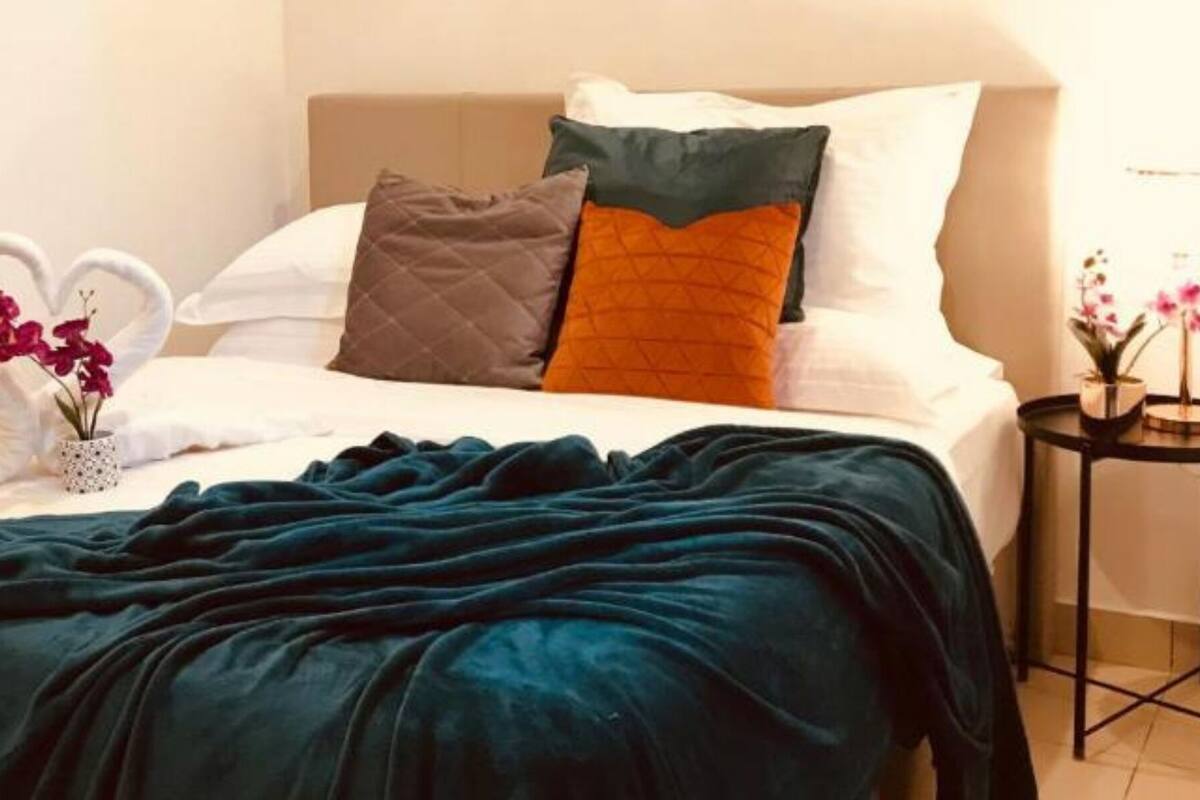 A neatly made bed with a heart-shaped towel and a vase of flowers, a dark green blanket, and a bedside table with a plant.