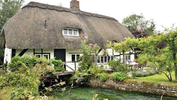 A traditional thatched-roof house with a small bridge over a waterway, surrounded by lush greenery and flowering plants.