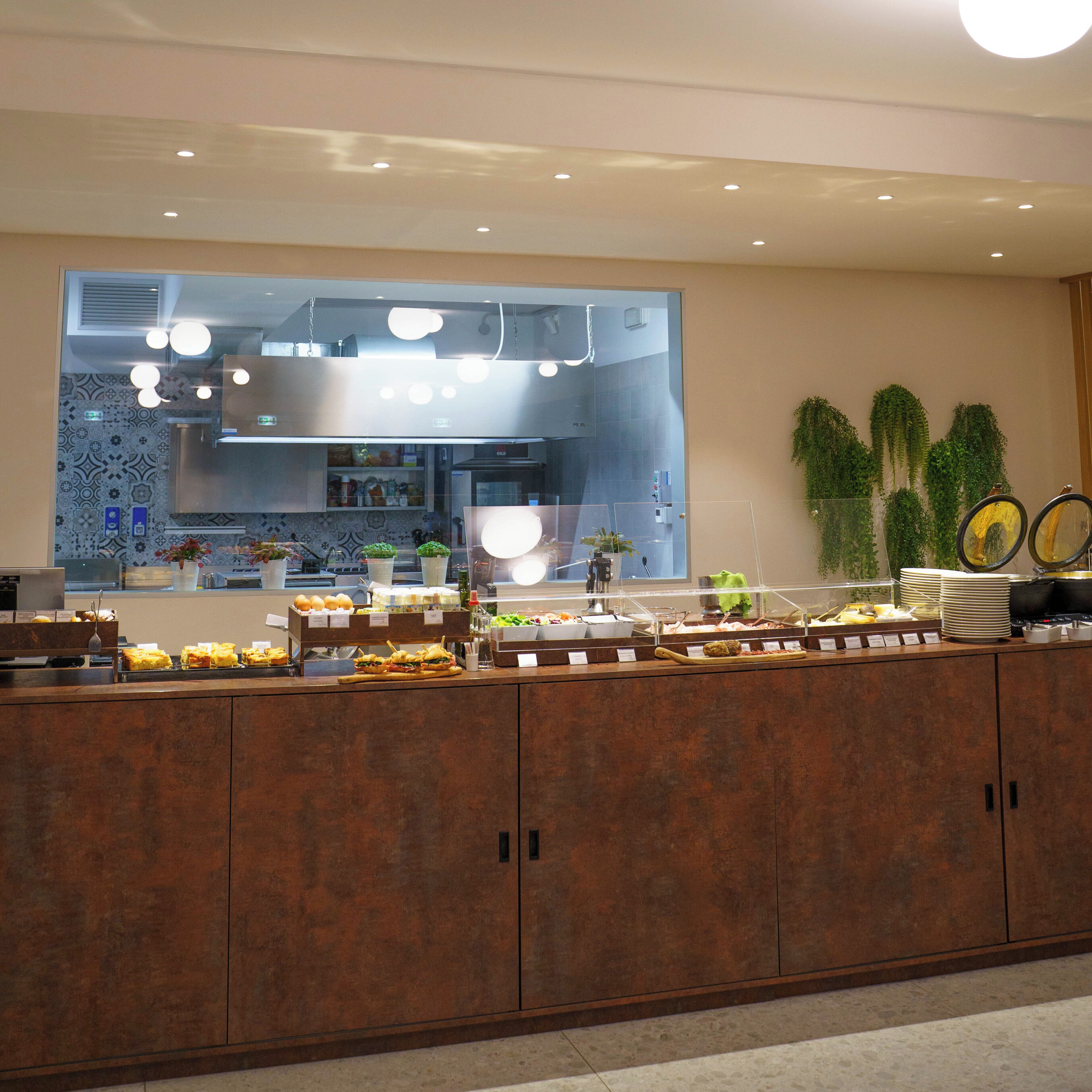 Un'area buffet con diversi cibi, una cucina sullo sfondo e una parete decorativa con piante.