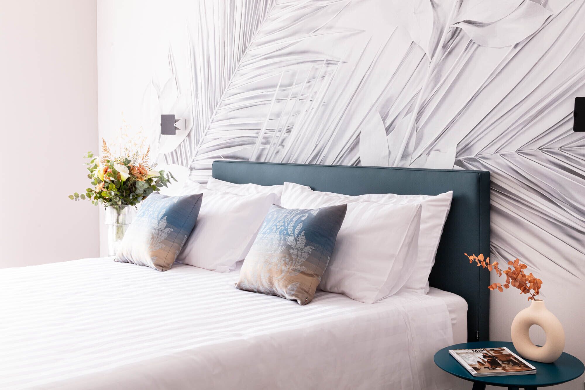 A neatly made bed with two pillows, a vase with flowers, and a magazine on a small table beside the bed.