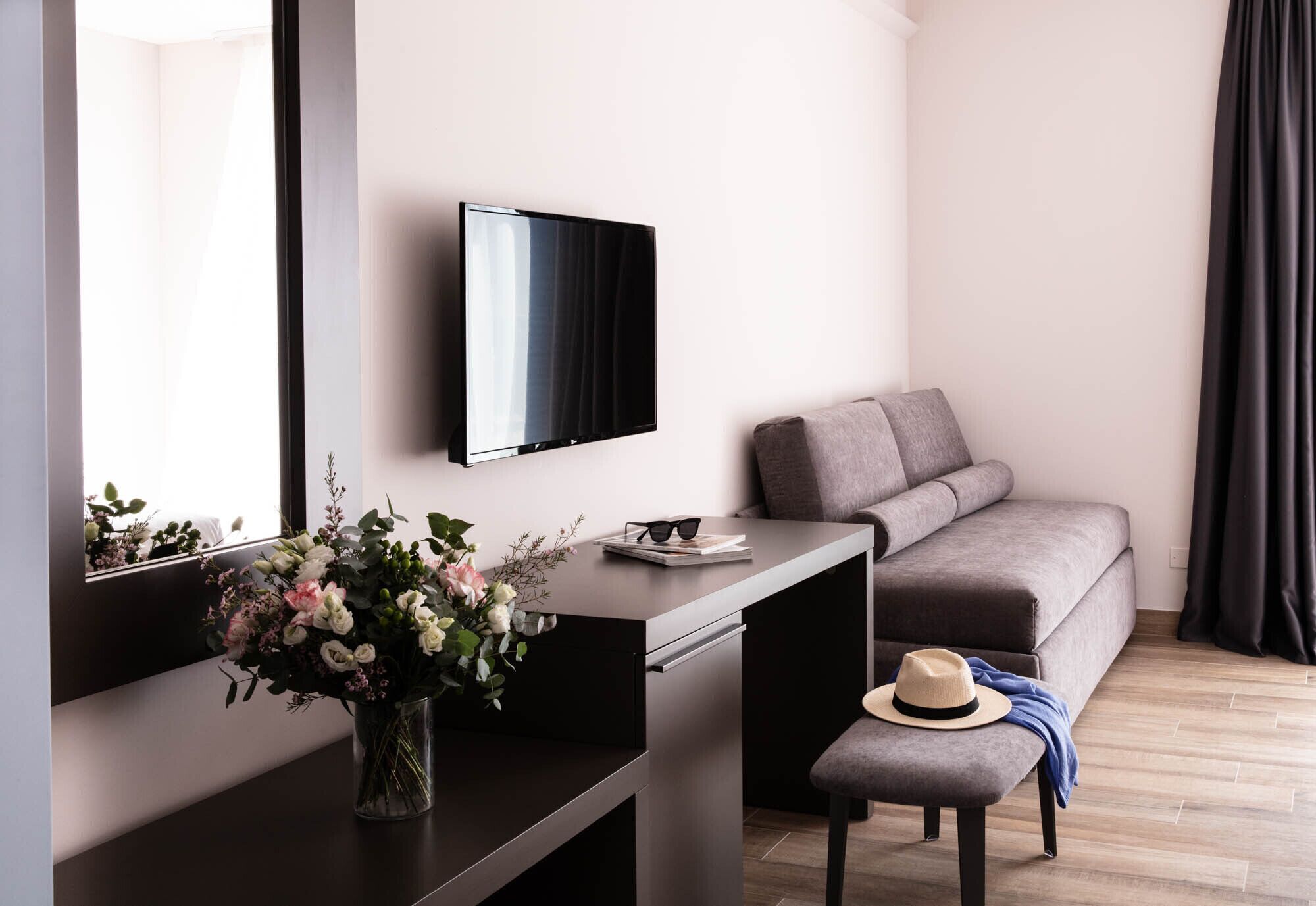 A modern hotel room with a flat-screen TV mounted on the wall, a grey sofa, a small table with a vase of flowers, and a wooden floor.