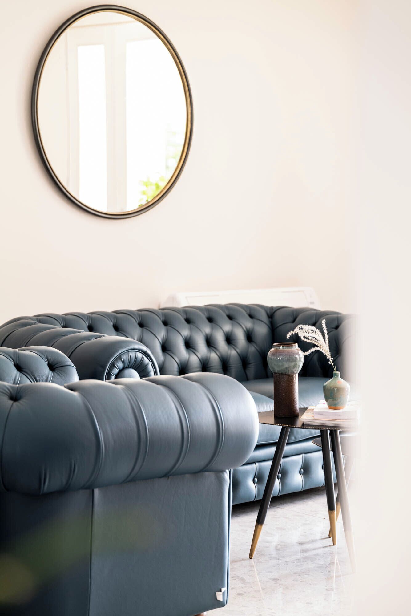 Un canapé bleu foncé à motifs capitonnés, avec un miroir rond au-dessus et une petite table à côté, sur laquelle se trouvent un vase et une plante.