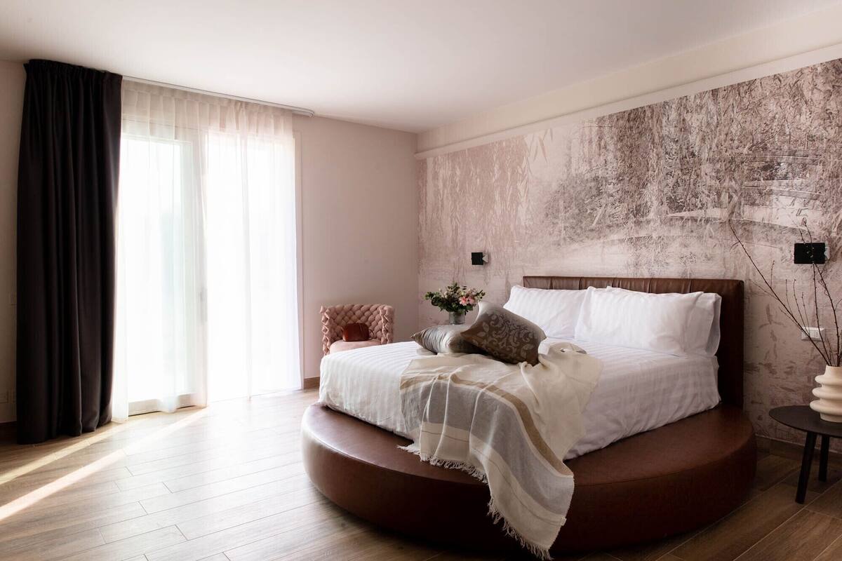 Una cama redonda con cabecera, una mesita de noche con un florero y un ventanal con cortinas translúcidas.