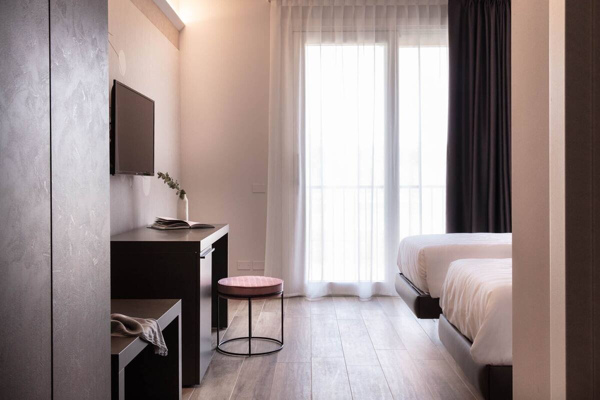 Una habitación de hotel moderna con cama, escritorio con silla, un taburete pequeño, un espejo y una ventana con cortinas.