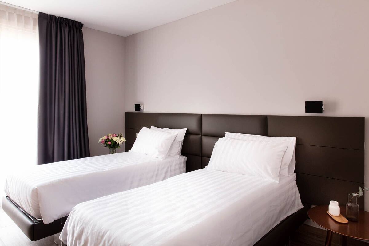 Habitación de hotel con dos camas individuales, un cabecero oscuro, una mesita de noche pequeña con un jarrón de flores y una ventana con cortina oscura.