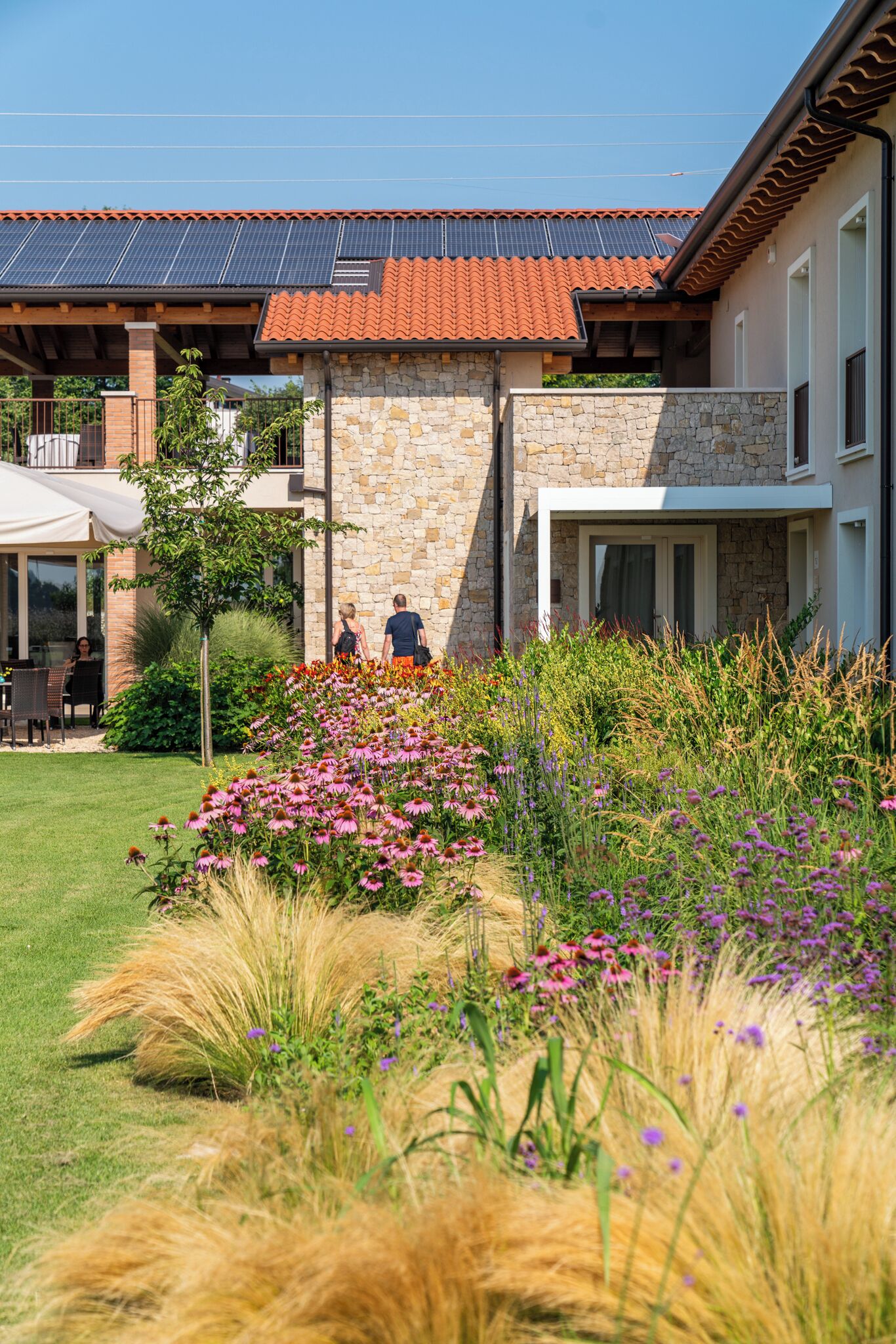 A modern house with a stone facade, a red-tiled roof, and a garden with various flowers and grasses.