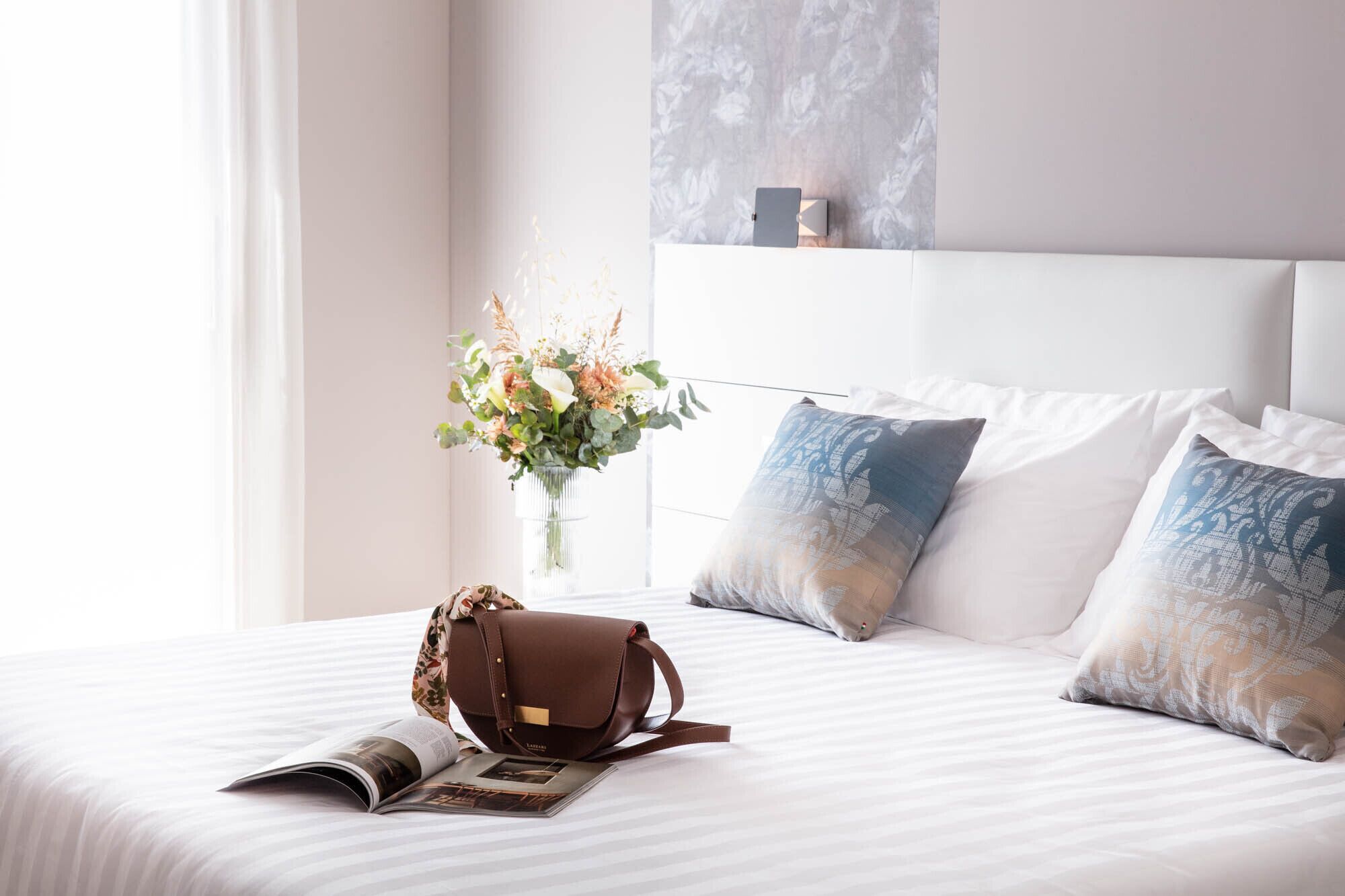 Un lit bien fait, avec un sac en cuir marron, un vase de fleurs et un magazine posés sur le lit.