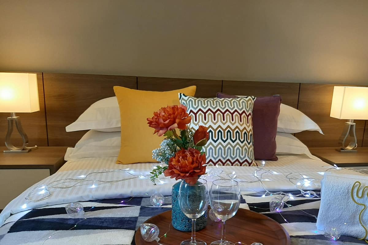 A neatly made bed with decorative pillows, a vase with flowers, and a tray with wine glasses and a towel.