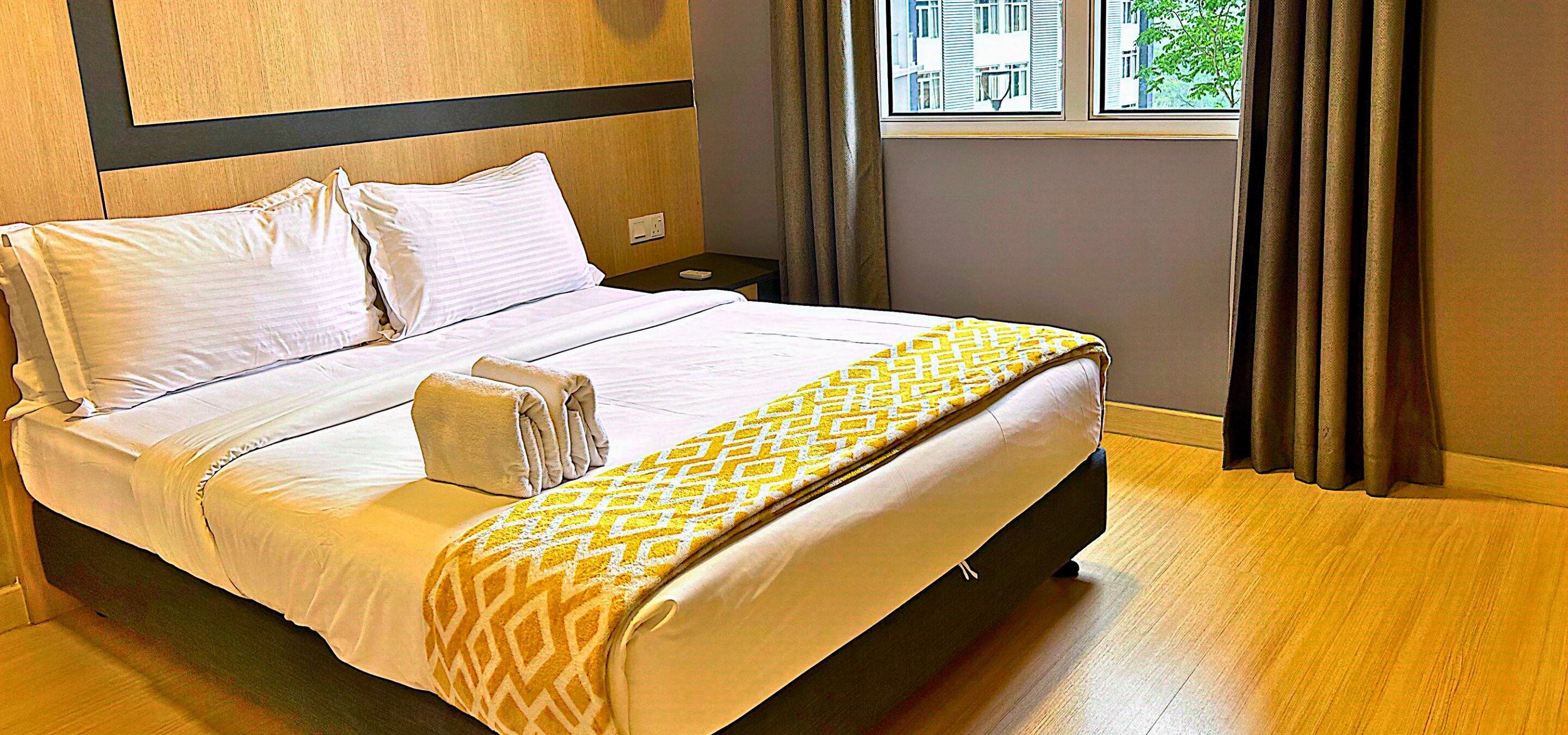 A neatly made bed with white linens and a patterned throw blanket, a bedside table with a lamp, and a window with curtains.