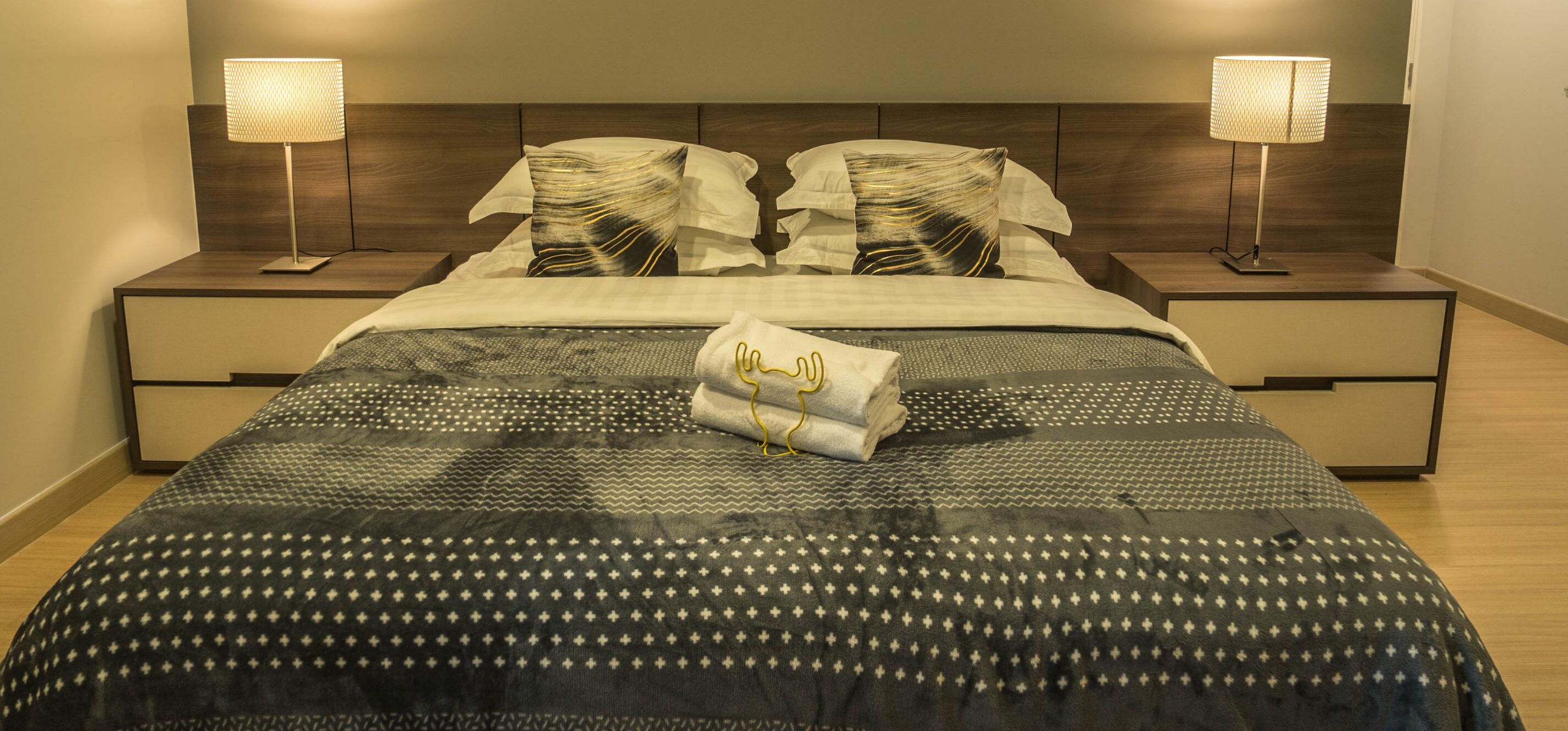 Una cama bien tendida con ropa de cama estampada, dos lámparas de noche y una toalla doblada sobre la cama.