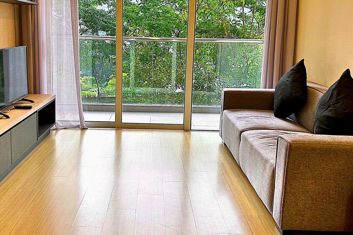 A modern living room with a flat-screen TV on a wooden stand, a sofa with black cushions, and a large window offering a view of greenery outside.