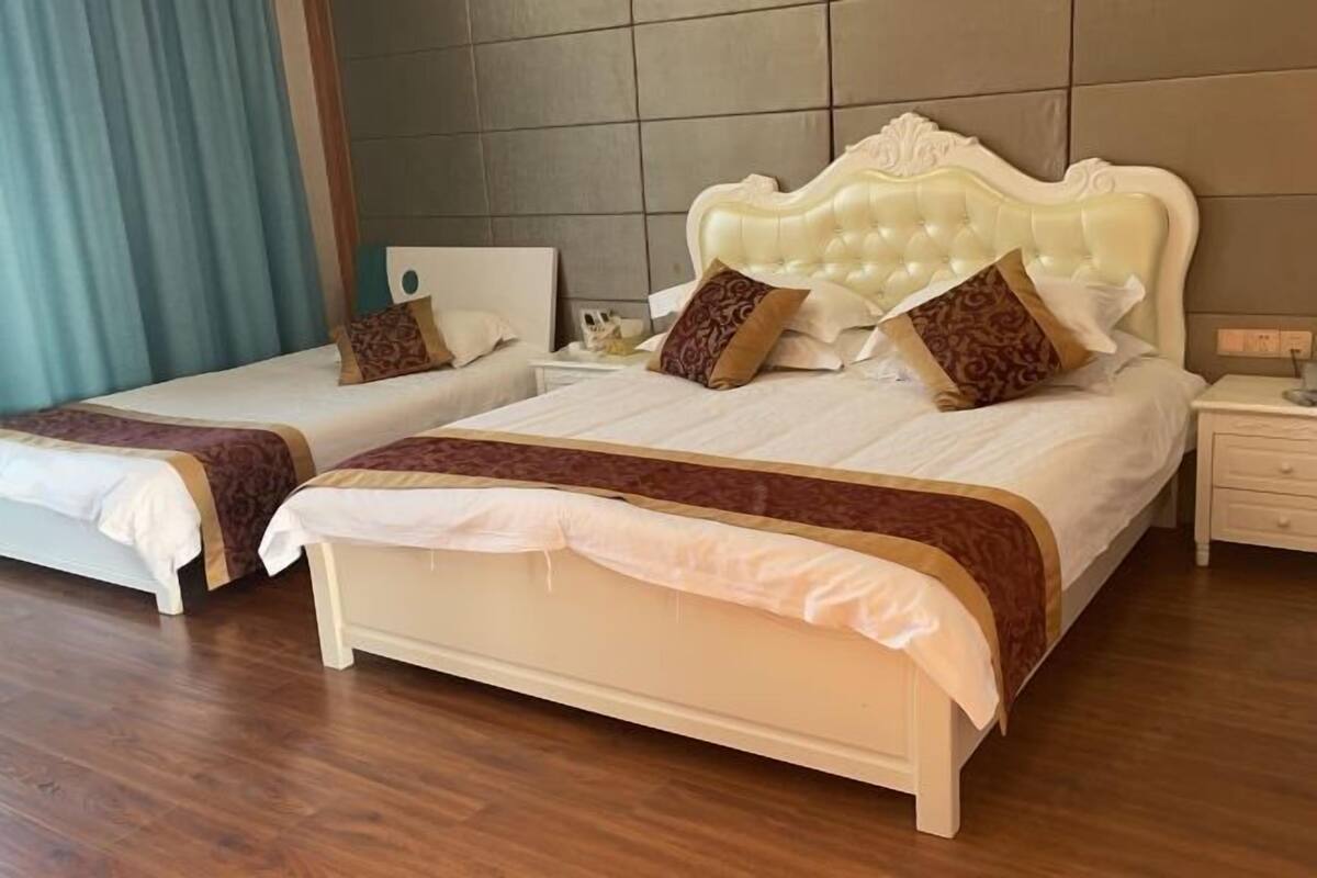 Duas camas de casal com cabeceiras iguais, uma mesa de cabeceira com abajur e piso de madeira.