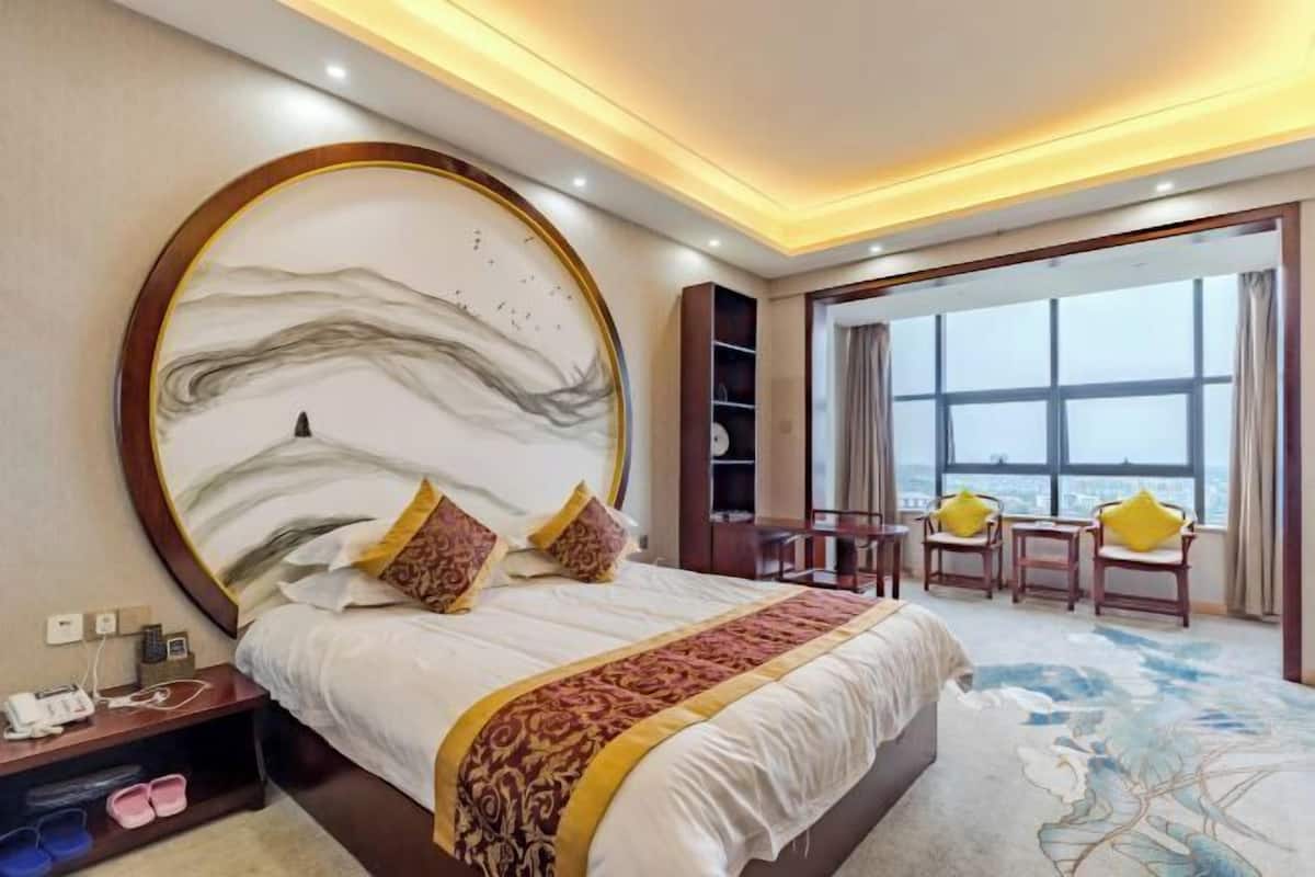 Quarto de hotel com uma cama grande, uma obra de arte circular na parede, vista através de uma janela ampla e um espaço de estar com cadeiras.