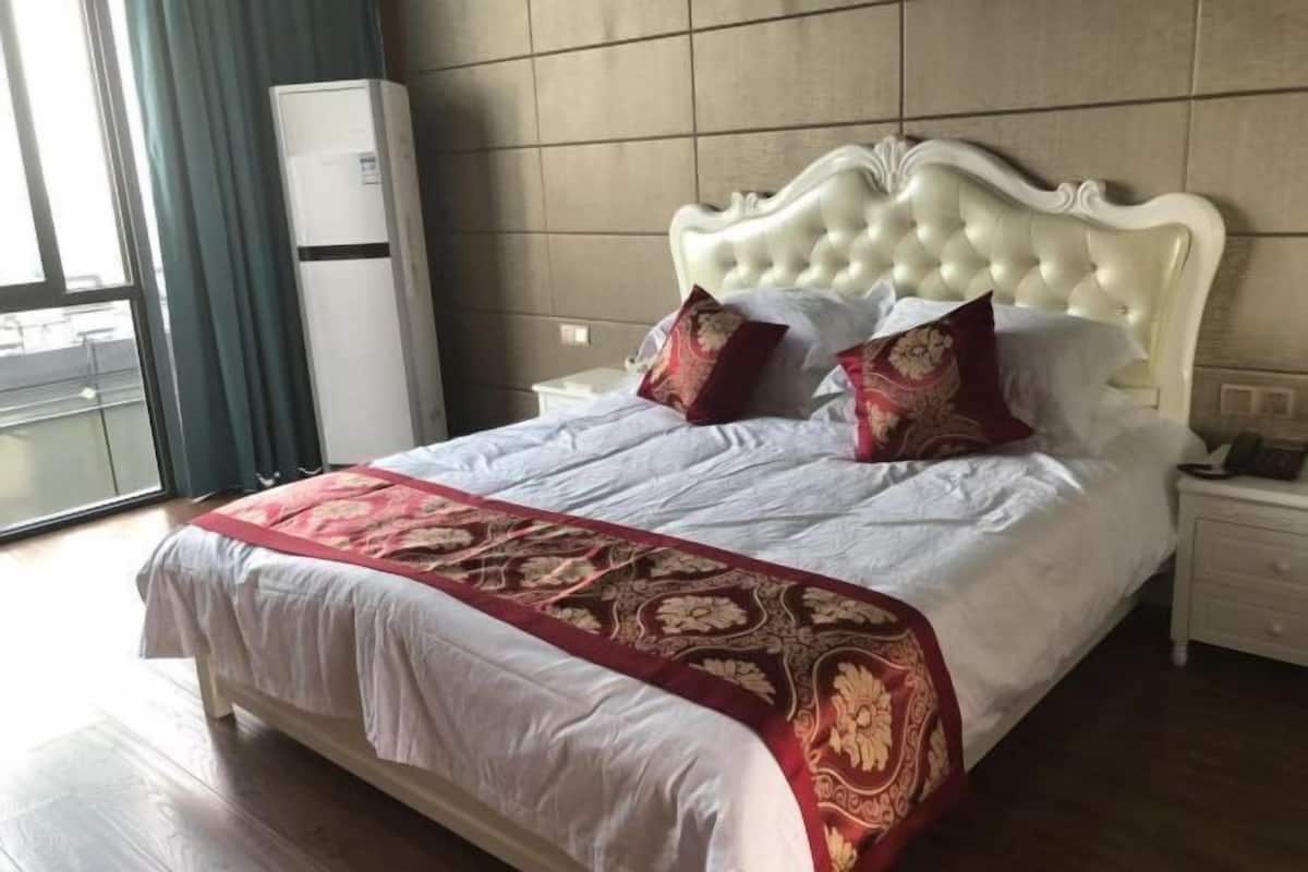Uma cama bem arrumada com cabeceira branca e colcha estampada em vermelho e dourado, uma mesa de cabeceira branca com abajur, e uma janela com cortinas.