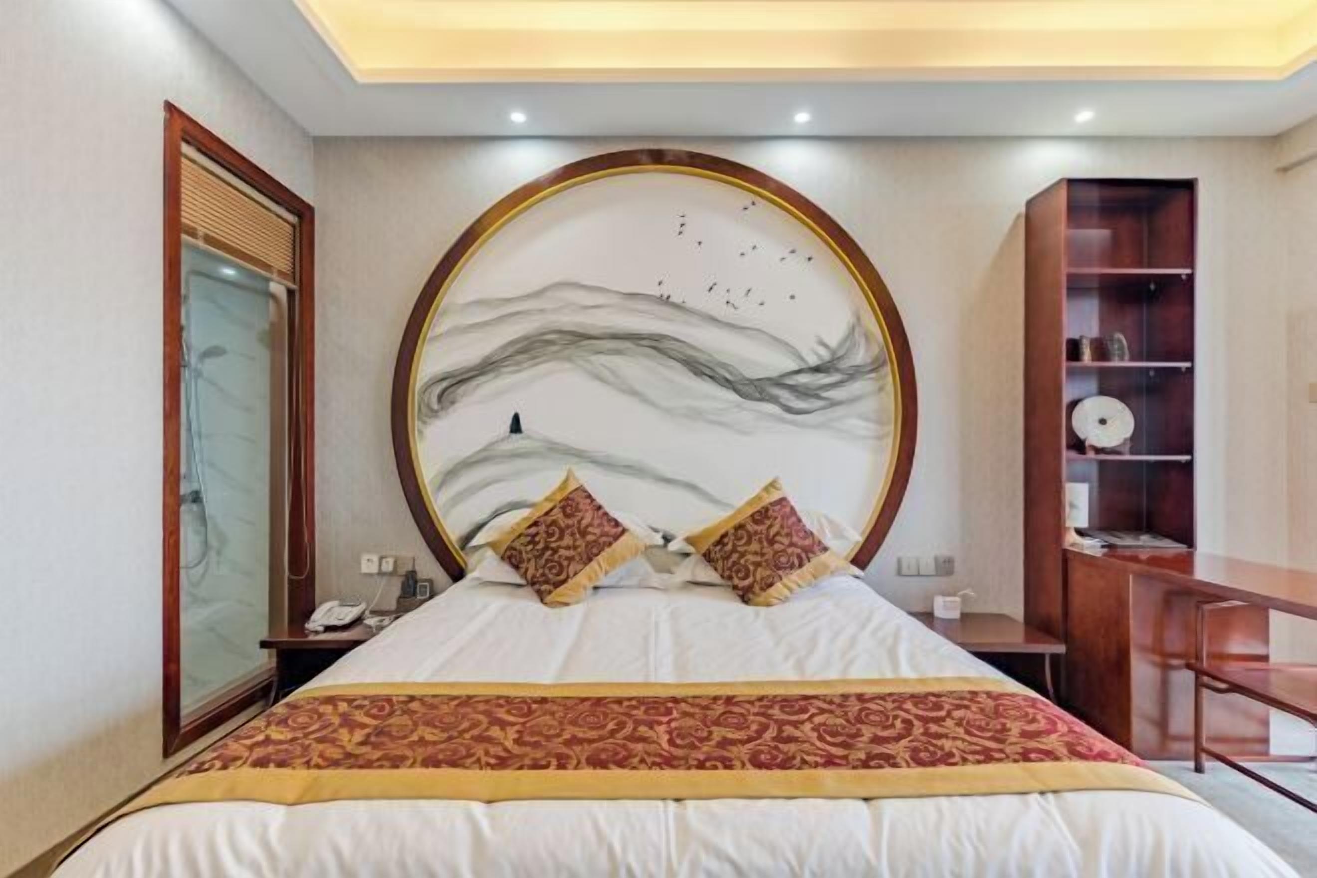 A hotel room with a large bed, a round headboard with a mural, a wooden desk, and a bookshelf.