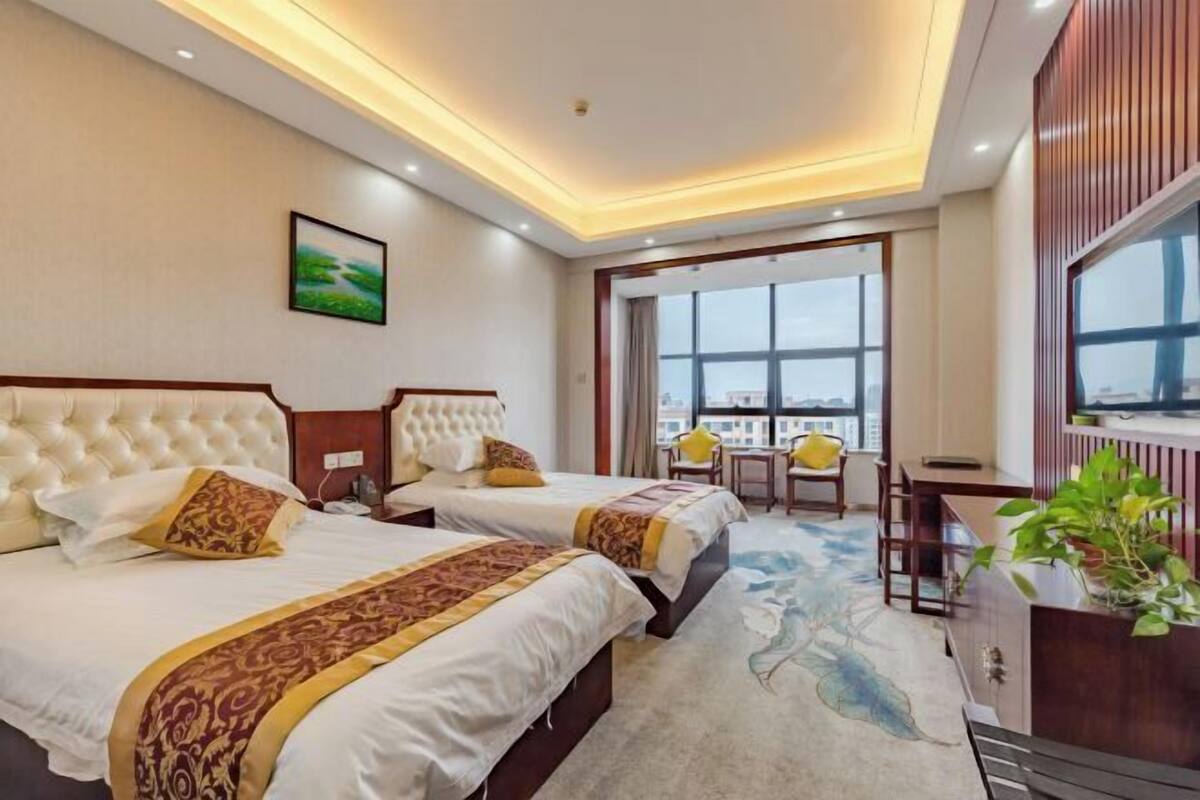 Quarto de hotel com duas camas, uma mesa, uma cadeira, janela com vista para a cidade e uma planta.