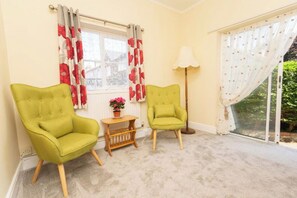 A room with two green armchairs, a small wooden table with a flower vase, a floor lamp, and a window with red and white curtains.