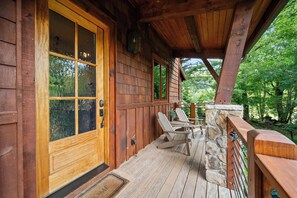 A wooden cabin with a large glass door, a porch with two Adirondack chairs, and a stone wall.