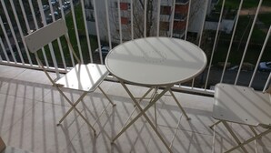 A balcony with a round table and two chairs, a railing, and a view of a parking area and buildings.