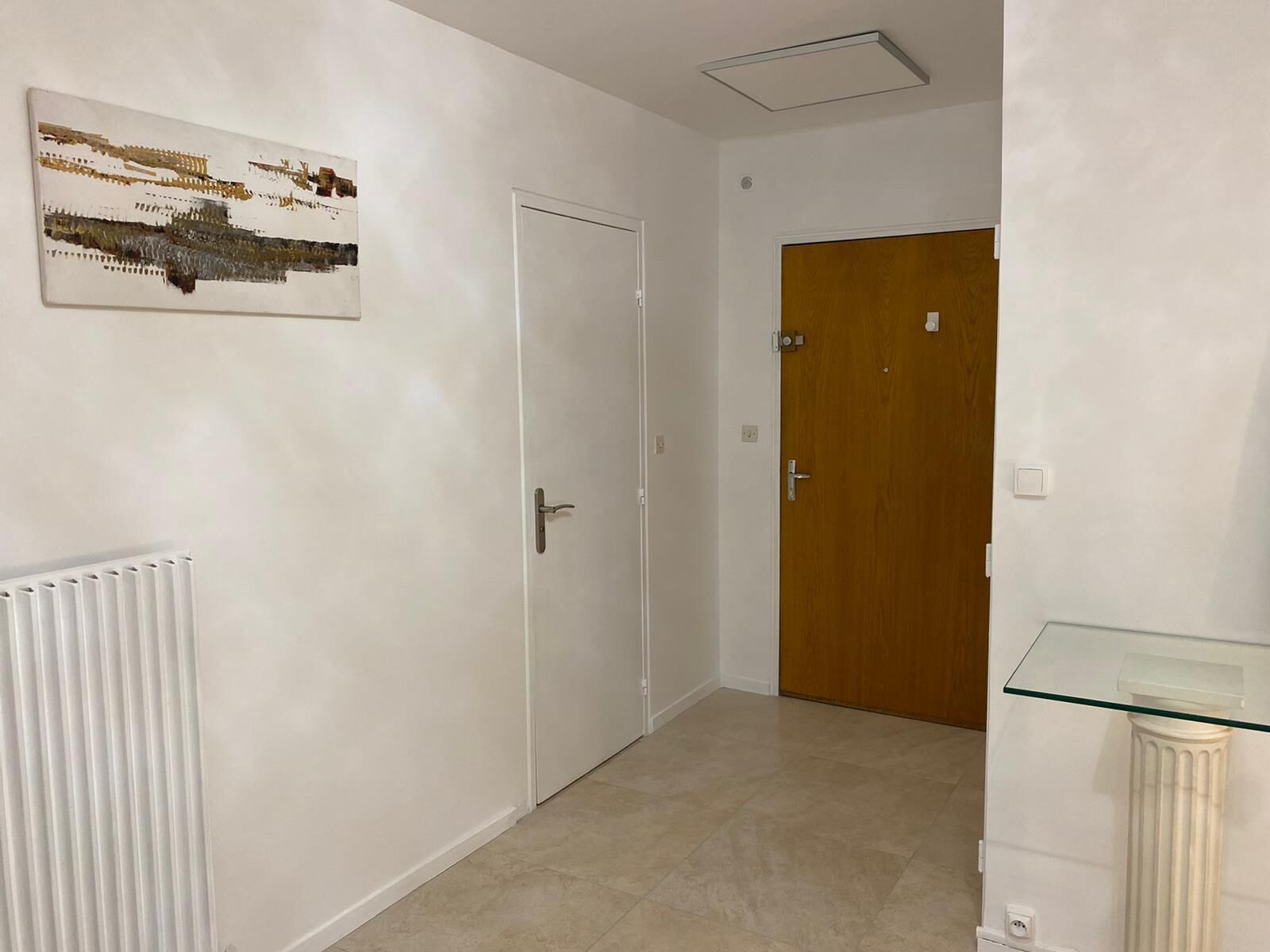 Corredor com paredes brancas, um radiador, uma mesa de vidro e duas portas.