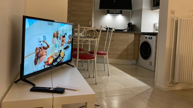 A modern living room with a flat-screen TV on a stand, a dining area with a metal chair and a table, and a kitchen area with a microwave and a washing machine.