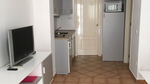 A compact kitchen with a microwave, a sink, and a small refrigerator. There is a TV on a white stand and a red folder on the counter. - Apartamento de 1 Dormitorio Cerca de la Playa (Fuengirola)