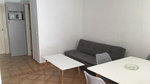 A compact living space with a gray sofa, a white dining table, and a small kitchen area including a microwave. - Apartamento de 1 Dormitorio Cerca de la Playa (Fuengirola)