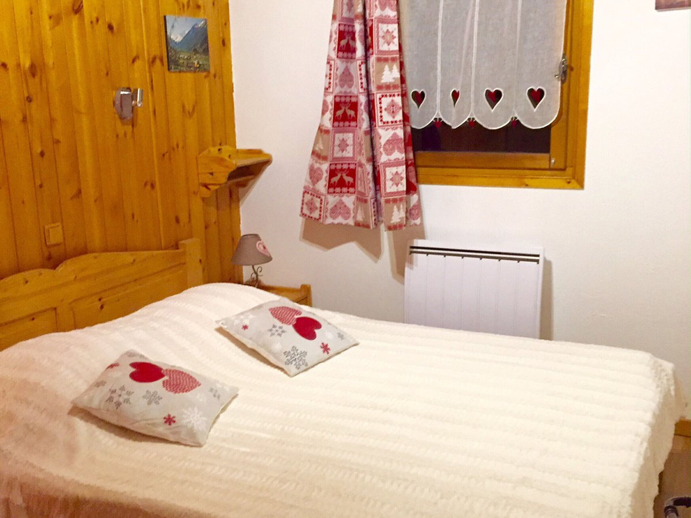 Une chambre confortable avec une tête de lit en bois, un lit avec une literie blanche et des oreillers à motifs de cœurs, une fenêtre avec un rideau blanc et un radiateur au mur.