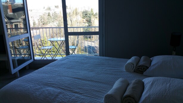 Habitación de hotel con una cama grande, un balcón con mesas y sillas, y vistas a edificios y árboles.