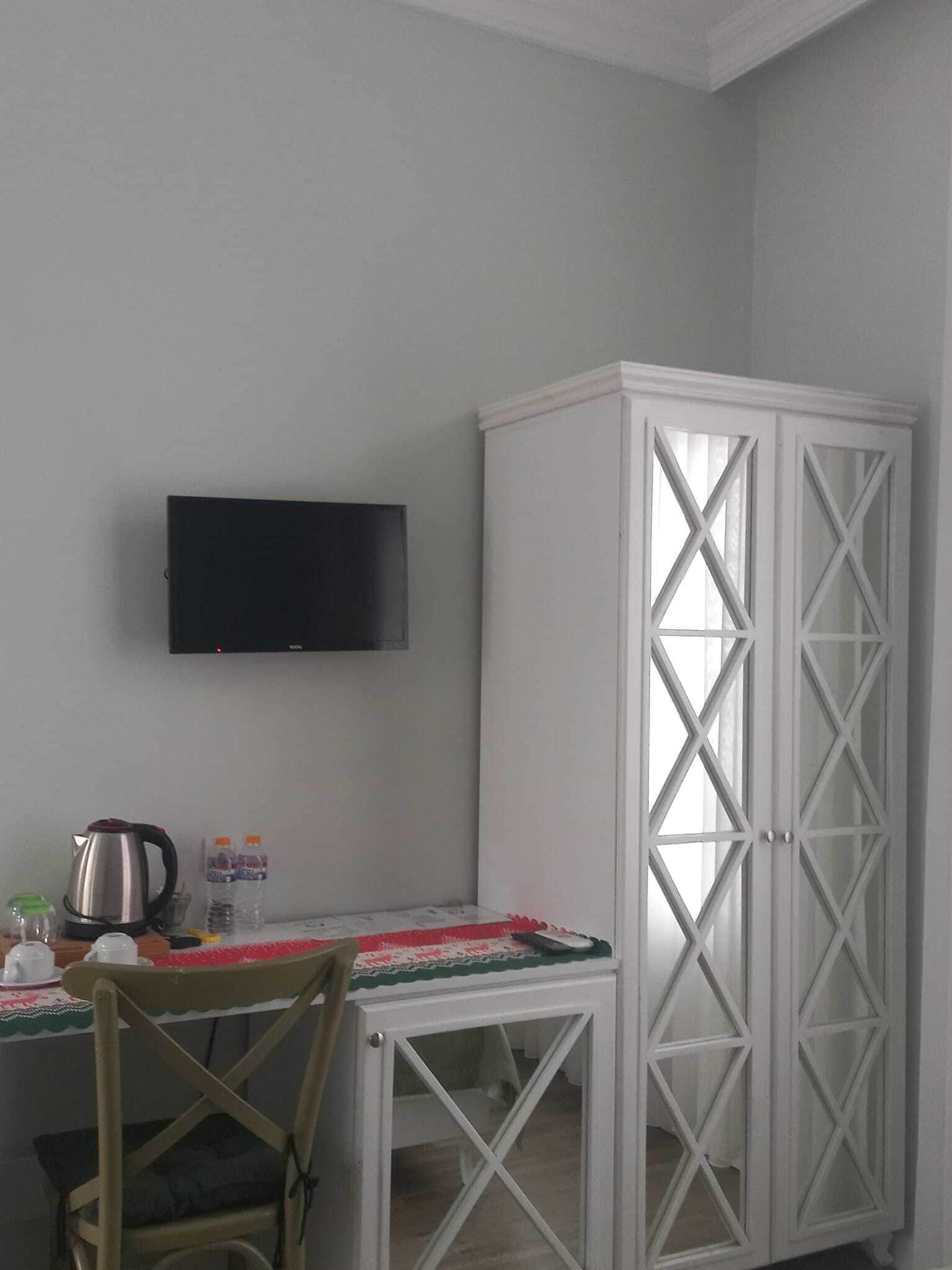 A room with a television mounted on the wall, a white cabinet with glass doors, a small table with a red and white tablecloth, and a chair.