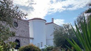 A building with a stone facade and a white upper section, surrounded by lush greenery and purple flowers. - Zarbalı Cunda (Ayvalik)