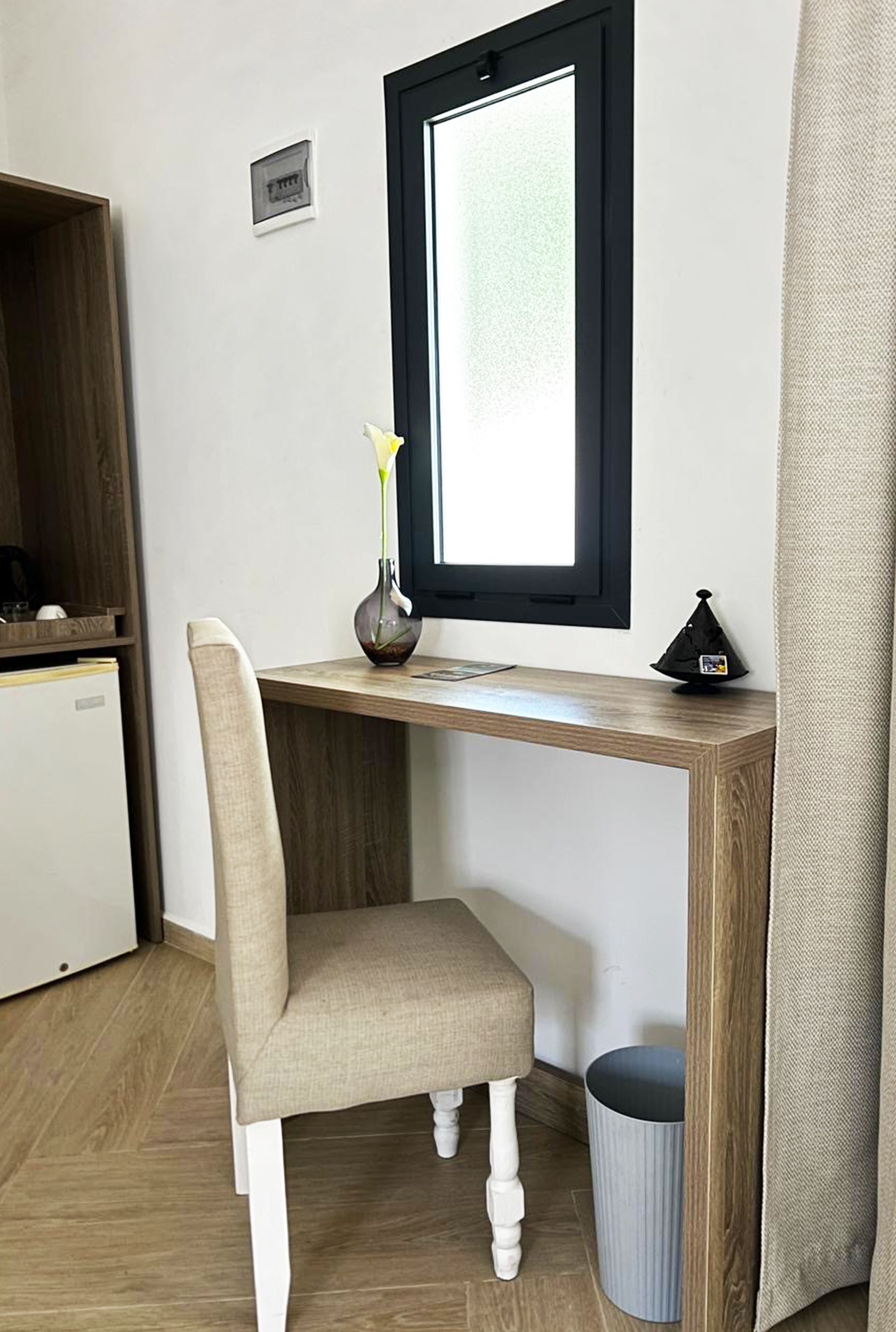 A corner desk with a chair, a small vase with a yellow flower, a black lamp, a window with a black frame, and a trash can.