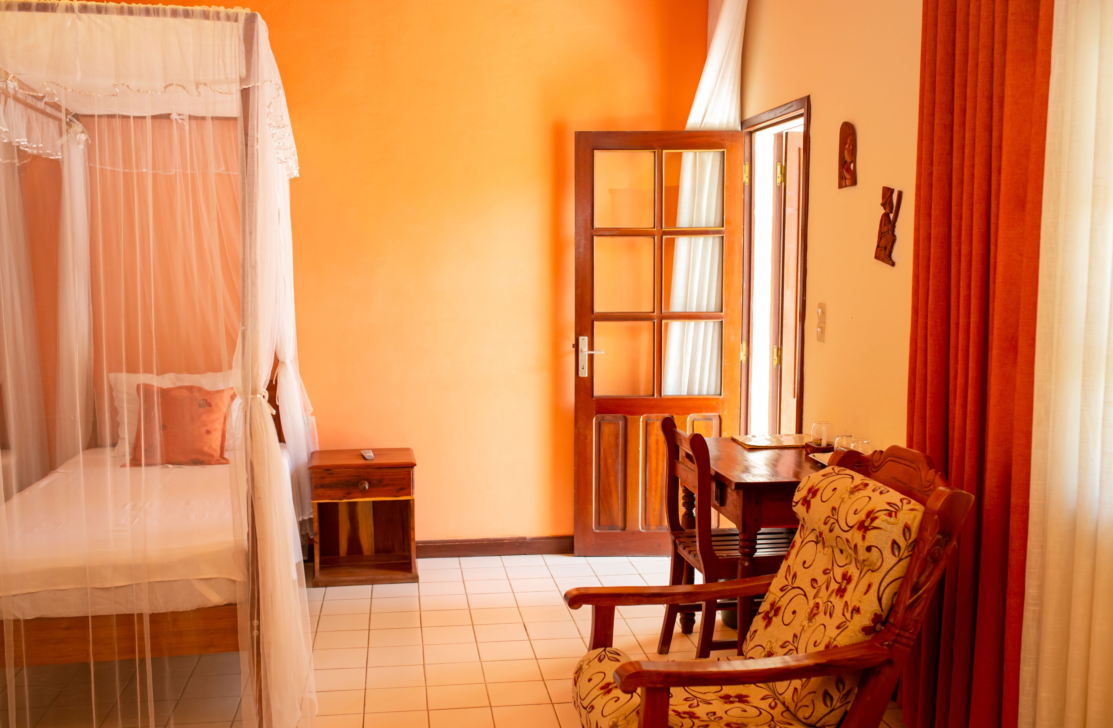 A room with a bed under a mosquito net, a wooden chair, a small table, and a door with a mirror.