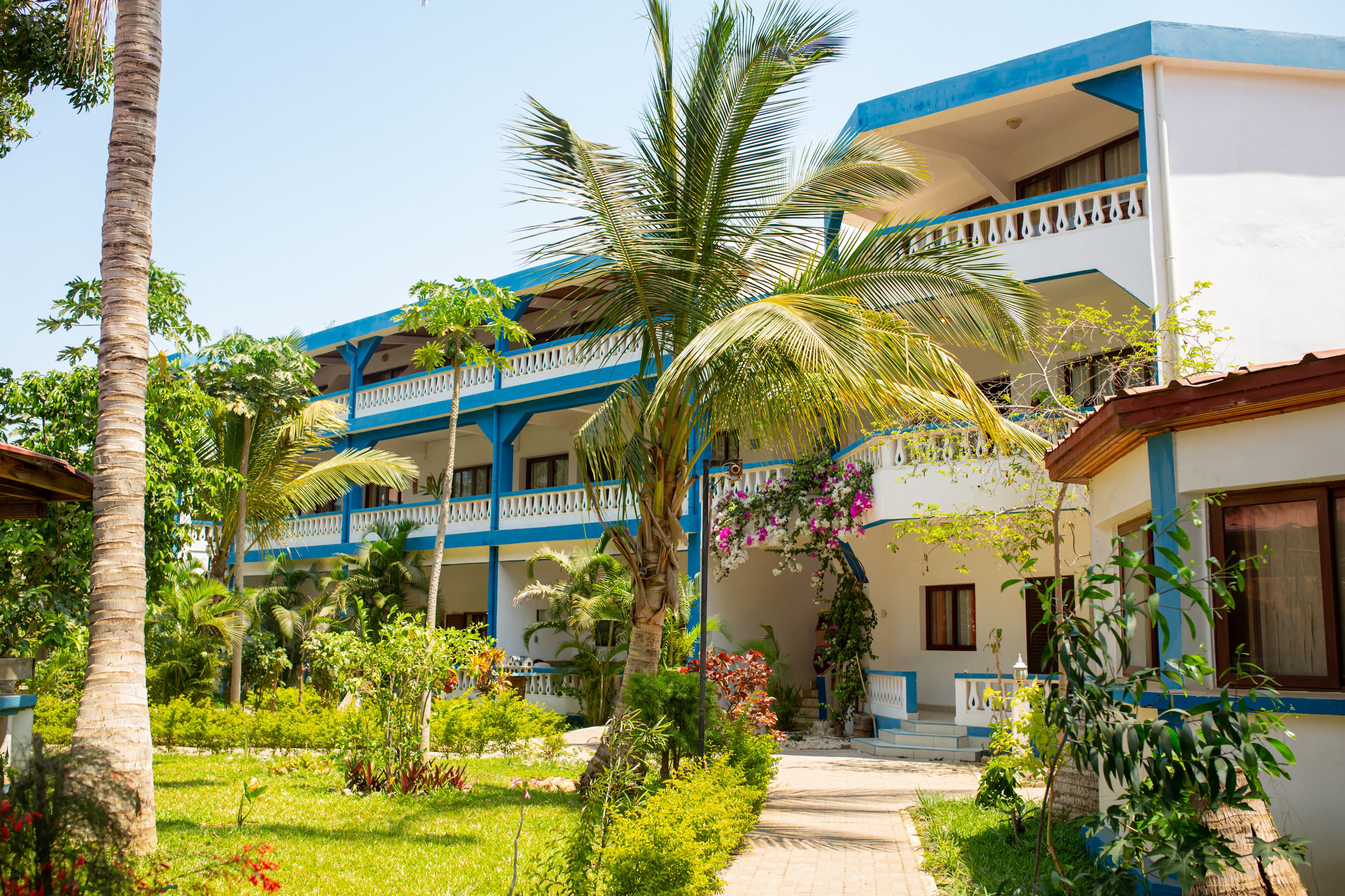 A hotel with multiple balconies, a garden with palm trees, and a pathway leading to the entrance.