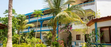 A hotel with multiple balconies, a garden with palm trees, and a pathway leading to the entrance.
