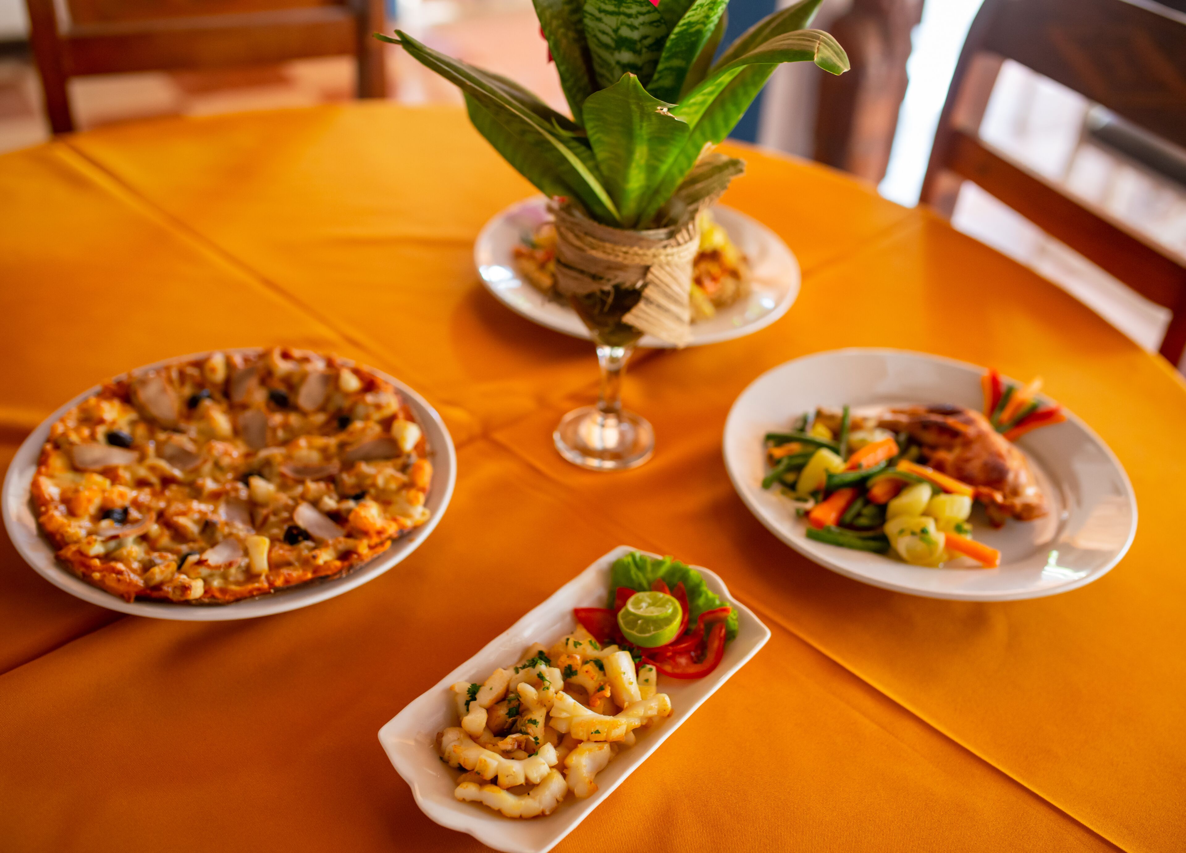 Una mesa con pizza, mariscos en un plato y un pequeño cuenco de pasta, todo sobre un mantel naranja.