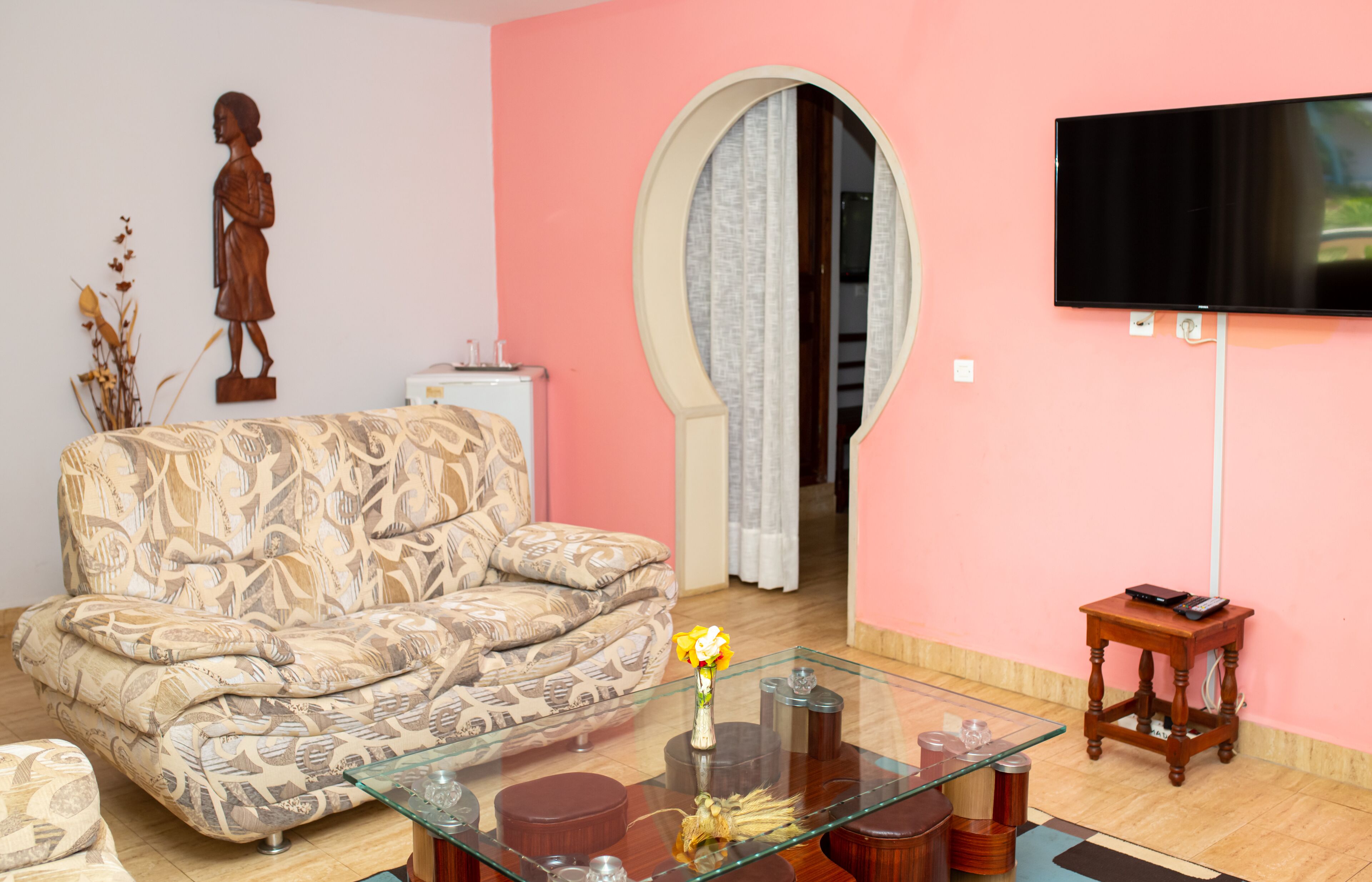 A living room with a patterned sofa, a glass coffee table with decorative items, a wooden side table, and a flat-screen TV mounted on the wall.
