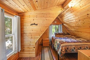 A wooden cabin bedroom with a sloped ceiling, a bed with patterned bedding, a window with curtains, and a small wooden nightstand. - Charming A Frame Cabin! Pet Friendly! Cozy 2 Bedroom! HOT TUB! Foosball Table! Wood Burning Fireplace! ½ Mile to Bear Mountain & Golf Course! (Big Bear Lake CA)