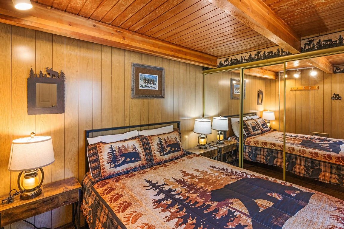 Une pièce aux murs recouverts de lambris en bois, un lit avec une couvre-lit à motifs forestiers, deux lampes de chevet et un miroir qui reflète le lit.