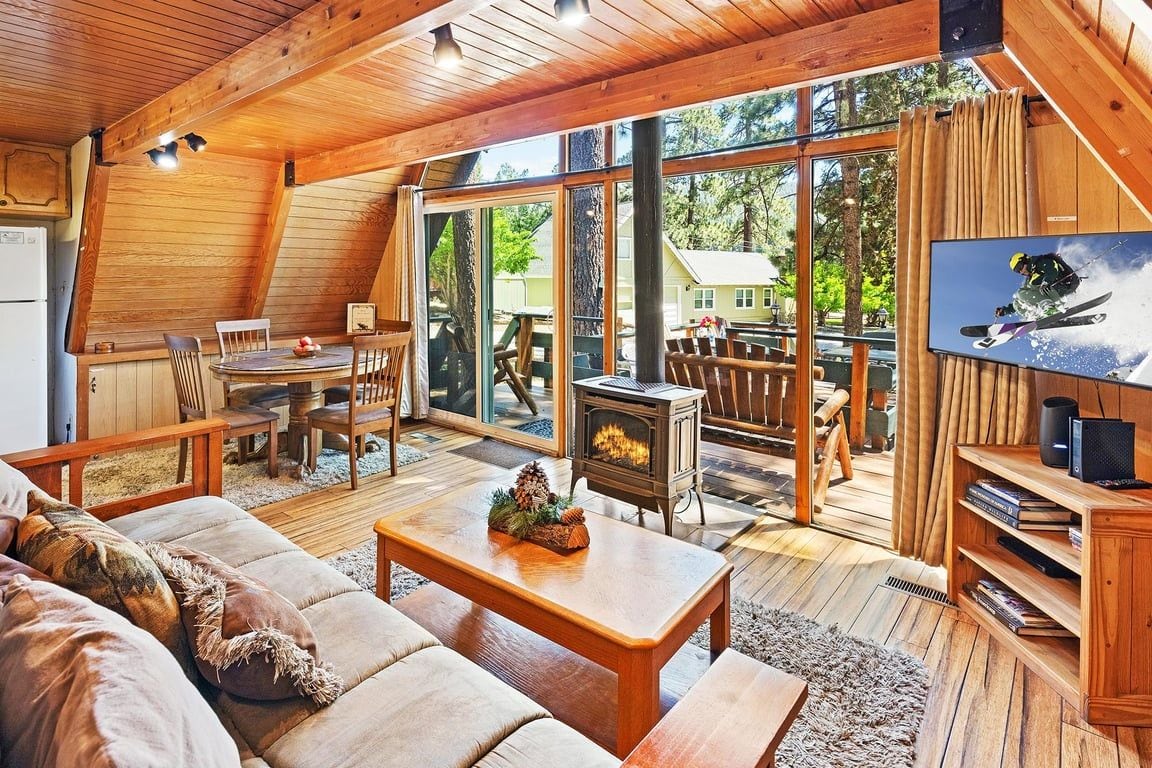A cozy cabin interior with a fireplace, wooden furniture, and a large window overlooking a deck.