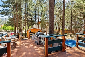 Een terras met buitenmeubilair, een barbecue en een hottub, omringd door hoge bomen.