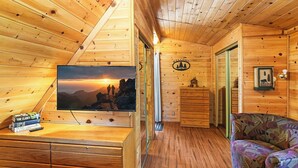 A cozy wooden cabin interior with a television displaying a scenic sunset, a wooden dresser, a patterned armchair, and a welcome sign on the wall.
