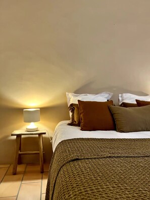 Un dormitorio con cama, almohadas, una mesita de noche con lámpara y suelo de baldosas.