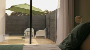 Un dormitorio con cama, almohada verde, silla blanca y un paraguas verde en el exterior.