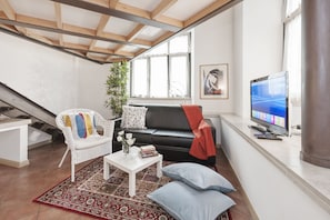 A modern living room with a black leather sofa, white wicker chair, and a red throw blanket. A flat-screen TV is mounted on the wall, and a large window provides natural light. - Inviting 3-bed Apartment in Roma 4mins to Colosseo (Rome)