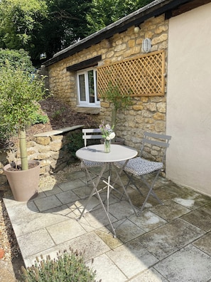 A stone building with a window, a wooden lattice, a potted tree, and a round table with patterned cushions. - Romantic Hideaway Cottage, Near Bridport, Dorset (Bridport)