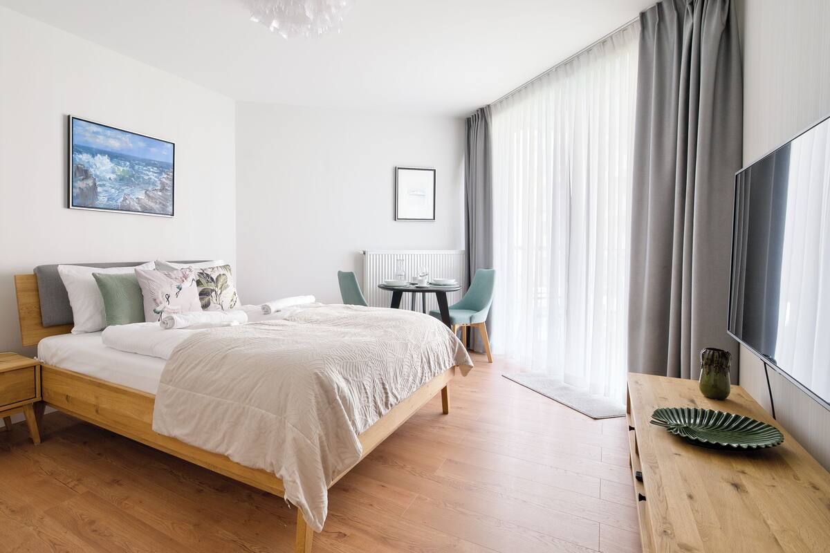 A modern hotel room with a wooden bed, a bedside table with a lamp, a small table with a chair, a large window with curtains, and a flat-screen TV.