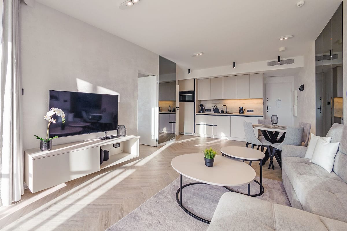 A modern living room with a flat-screen TV, a dining area with a round table, and a kitchen in the background.