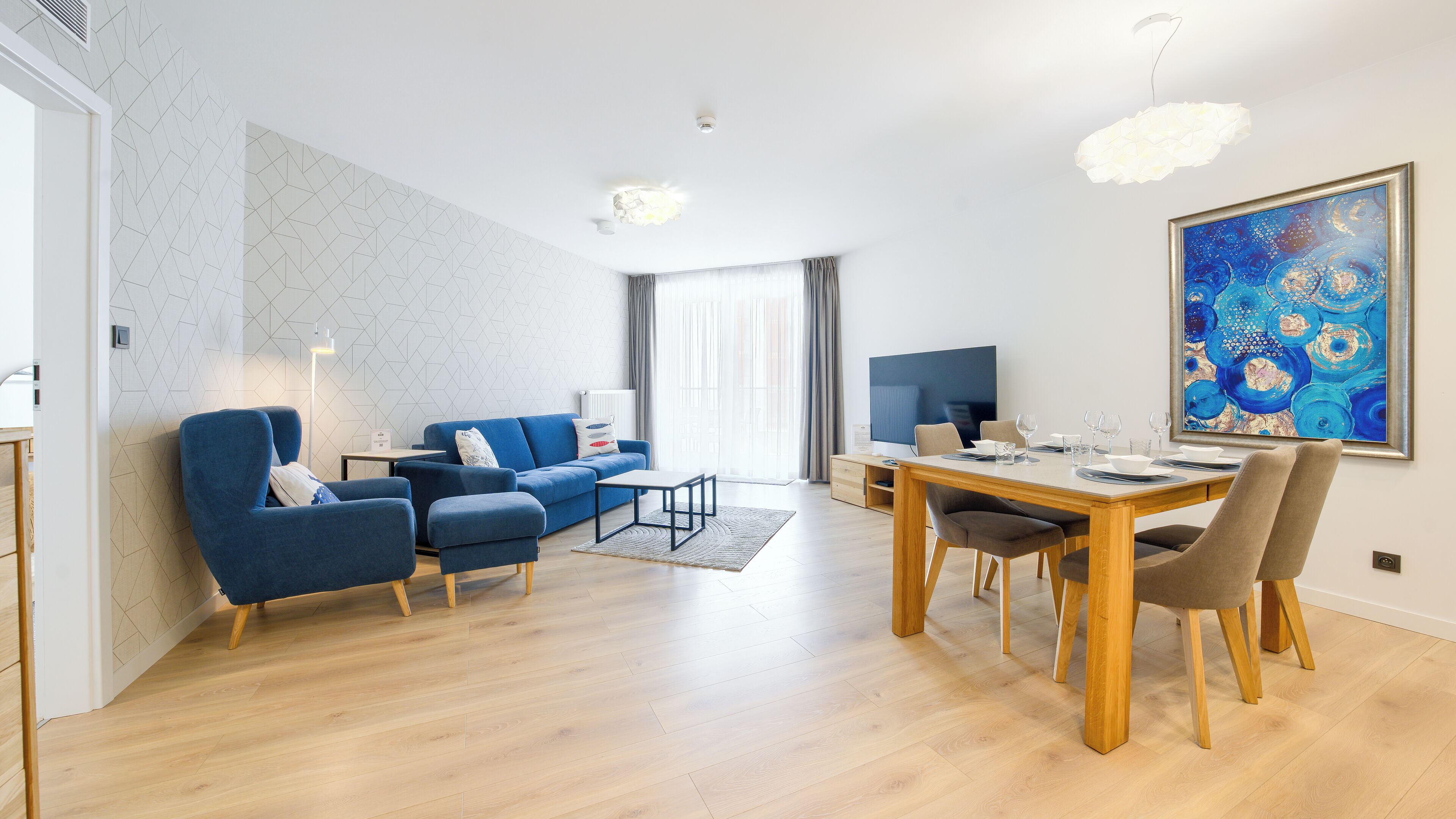 Un salón moderno con un sofá azul, una mesa de comedor preparada para cuatro personas y un cuadro abstracto grande en la pared.